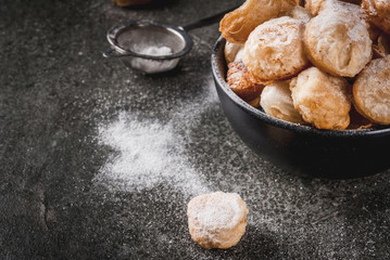 Homemade baking, puff pastries. Trendy food. Cronuts popcorn, puff donuts holes, in a black bowl and a paper bag, with powdered sugar. On a dark stone table. Copy space