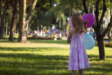 Little girl in pink dress with balloons in his hand standing on the grass on a background of trees