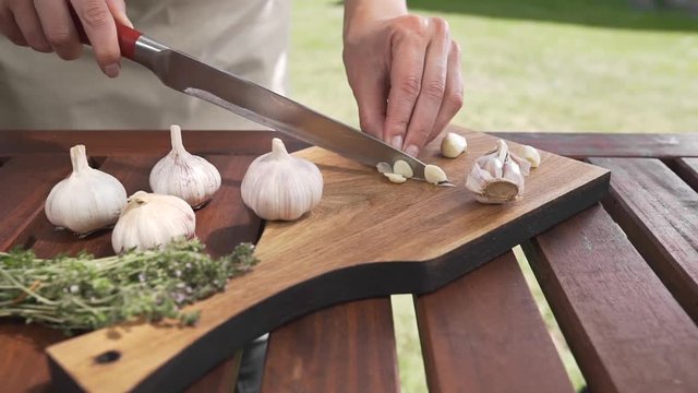 The cook slices garlic on the wooden board by sharp knife outside, cooking outside, cooking healthy food, summer meals