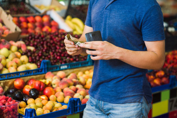 Young man paying cash at food market. Cash payment in close-up. Fresh fruits in the background.