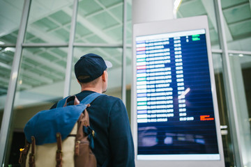 A young male traveler with a backpack in casual style looks at the information board at the airport. Getting information about the flight. Transfer.
