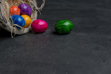 multi-colored painted eggs scattered from a straw nest against a dark gray stone background. Easter eggs. Happy Easter frame. Copy space.