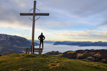Obraz premium Lonely hiker standing at a summit cross at Wandberg mountain in Chiemgau Alps mountain range in Austria shortly after sunrise. Tranquil scene with fog in the lowland. The cross is a christian symbol.
