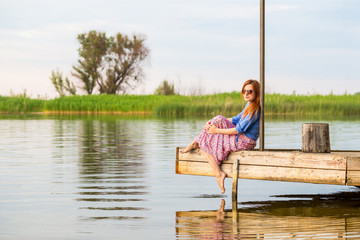 Beautiful young red woman sitting on a wooden pier in a colorful long sarafan dress and putting her...