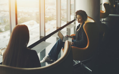 Business meeting two woman: caucasian and afro american with laptop and curly hair, both are sitting in armchairs on high floor of skyscraper near window, office luxury interior with reflections