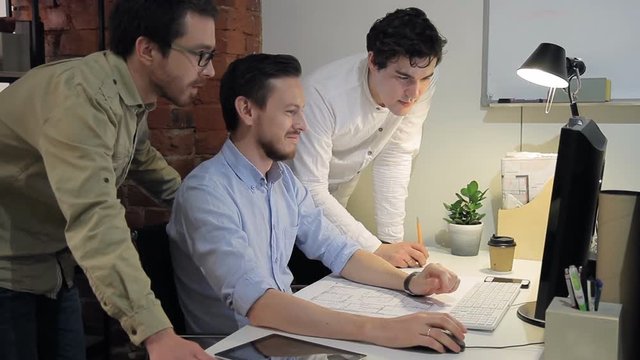 team group work on computer on architecture building project inside office. Man in blue shirt sit in middle, speak, explain working on drawing, show result on screen. man with beard stand compares