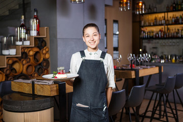 Ganson, officers, beautiful deushka with a smile and gathered hair in working form apron accepts an order in restaurant standing near the servered wooden tables