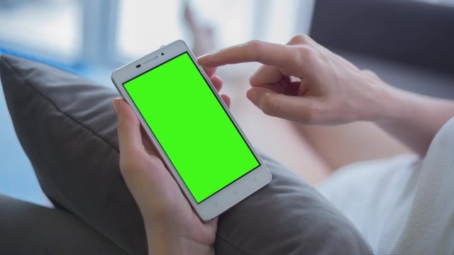 Young Woman in white top sitting on sofa uses smartphone with pre-keyed green screen. Few types of gestures - scrolling up and down, tapping, zoom in and out. Perfect for screen compositing
