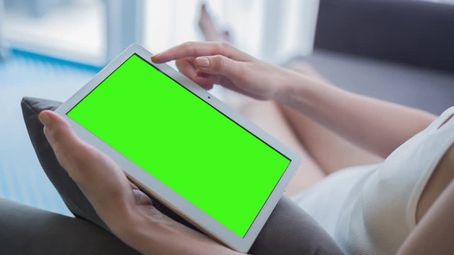 Young Woman in white top sitting on sofa uses Tablet PC with pre-keyed green screen. Few types of gestures - scrolling up and down, tapping, zoom in and out. Perfect for screen compositing