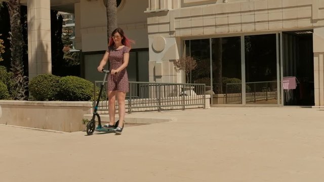 Young woman rides scooter in summer day outdoors. She moves on two-wheeled compact transport along smooth surface, pushing one foot and firmly holding onto t-shape steering wheel. Spectacular brunette