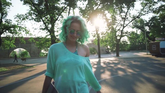 Portrait of young stylish hipster girl with green hair walking with long board during sunrise, slow motion, gimbal shot