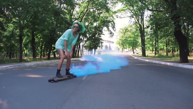 Beautiful young girl with green hair riding on longboard with blue smoke in park, slow motion, tracking shot