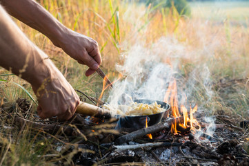 Cooking food in pan on camping fire. Hands hold frying pan and fork preparing simple tourist food...