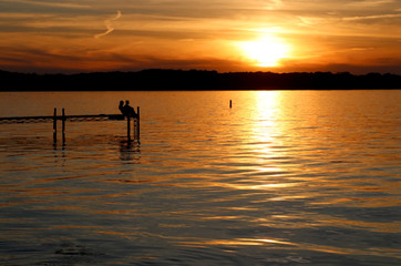 Fototapeta premium Summer sunset over the lake. Landscape with golden sunset and silhouettes of people enjoying the beautiful evening on a lake Mendota pier in the city of Madison, Wisconsin, USA.