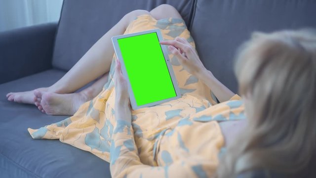 Young Woman in yellow dress laying on couch uses Tablet PC with pre-keyed green screen. Few types of gestures - scrolling up and down, tapping, zoom in and out. Perfect for screen compositing