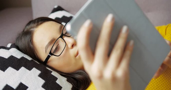 Attractive young woman scrolling pages, phtos on tablet device while lying on the couch. Beautiful woman in glasses typing on tablet computer and smilling at home. Close up