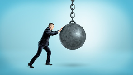 A businessman on blue background pushing at a hanging unmoving wrecking ball.