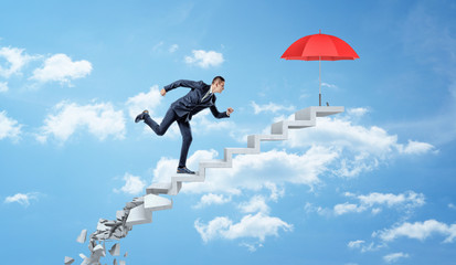 A businessman running up on crumbling concrete steps through the clouds to reach a red umbrella.