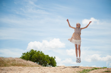 happy cute little girl wearing summer dress jumping outdoors on sky and tree background