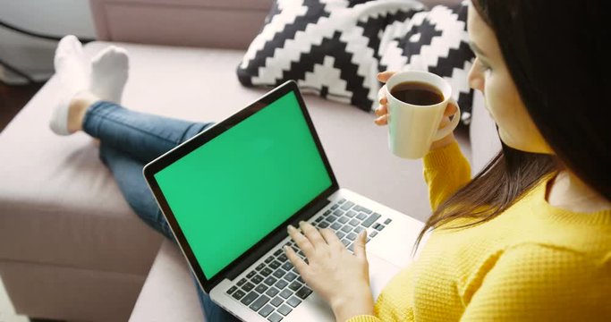 Young woman using laptop pc with green screen, drinking coffee while sitting on the couch in the living room. Back view. Chroma key