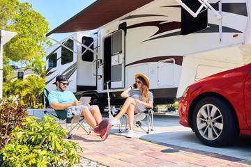 Couple with electronic devices sits on chairs near camping trailer