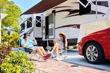 Young couple sits near camping trailer,smiling.Men talks on mobile phone , woman uses electronic device on chair near red car and green palms.Family spending time together on vacation in rv park