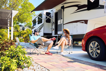 Young couple sits near camping trailer,smiling.Woman and men drinking water, relaxing on chairs near car and palms.Family spending time together on vacation near sea or ocean in modern rv park