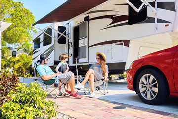 Young family sits on chairs near camping trailer and car