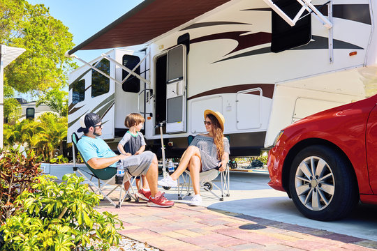 Young family sits on chairs near camping trailer and car