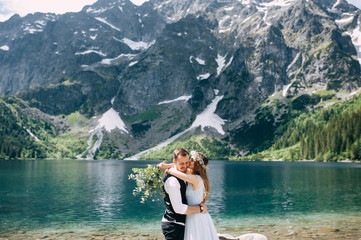 bride with the beautiful blue dress and groom hugging with views of the beautiful green mountains and lake with blue water