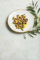 Pickled green Mediterranean olives in virgin olive oil on white ceramic plate and olive tree branch over grey marble background, selective focus, copy space, vertical composition