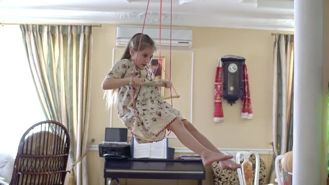 Little girl in linen dress hanging on trapeze at home and doing tricks