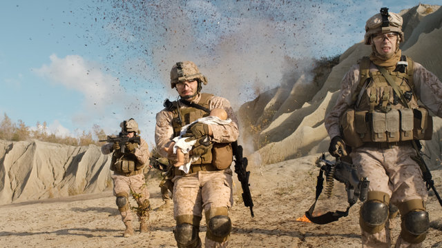 Soldier Running away from Explosions behind Carrying a Baby. While other Members of the Squad Covering Them During Battle in the Desert.