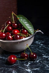 Several red sweet cherries and big green leaf on the table. Fresh organic cherry in colander on dark marble background..