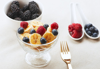 Tasty breakfast of bowl with banana, blueberries and raspberries along with napkin and fork and a bowl with blackberries.
