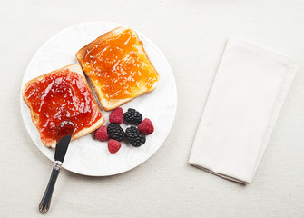 Top view of tasty toast breakfast with strawberry and peach jam along with blackberries and raspberries, and a napkin.