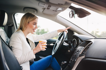 Beautiful young blonde woman driving a black car and drinking tea. Close up portrait. Smiling girl.