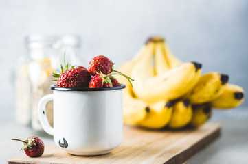 Fresh strawberries in a white metal cup, bananas on cutting board and cereals  over gray concrete background