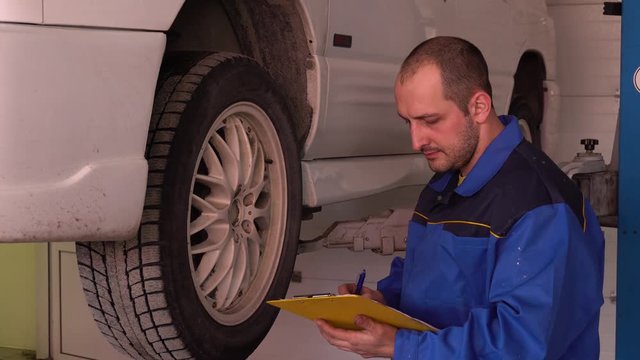 Car mechanic in blue robe inspects the rubber and writes to the tablet close-up.
