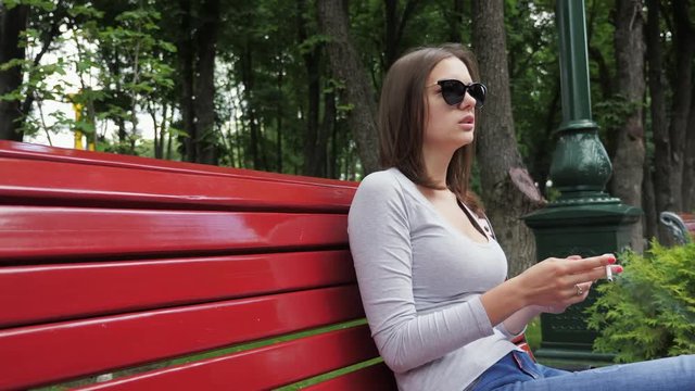 Young beautiful girl in sunglasses smoking a cigarette outdoors in a park bench
