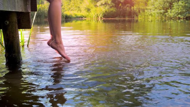 Woman sitting on the edge of a wooden jetty at lake, feet near the water surface, slow motion.
