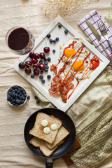 English breakfast - fried egg, bacon, bread blueberry.and cherry in frying pan on wooden board over White tablecloth background