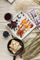 English breakfast - fried egg, bacon, bread blueberry.and cherry in frying pan on wooden board over White tablecloth background