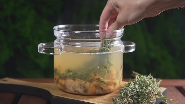 The cook puts bunch of thyme to the hot shrimp broth in the glass pan standing on the wooden board outdoors, seafood meal, seafood soup, first course, asian cuisine, food cooking outside
