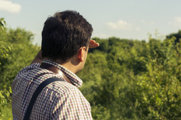 A young man in a shirt looks into the distance with his hand to his face against the background of blue sky and green trees