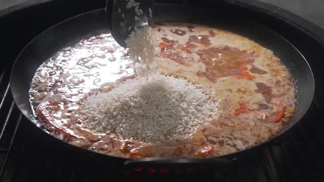 The cook makes paella on the open fire and adds rice to the other ingredients in the big hot pan, grill anf barbecue, spanish food, paella with seafood, food cooking outdoors