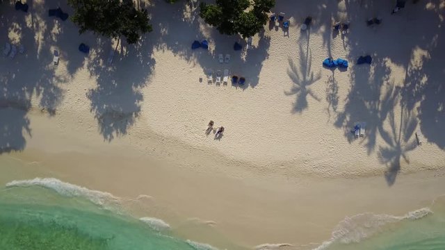 Aerial drone camera ascending and rotating high above the couple in love lying in the sun on beach bed deck chairs and drinking cocktails at empty tropical white sand beach on atoll island resort with