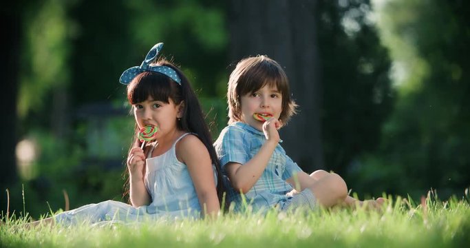 two little children with a Lollipop,Caucasian boy and Asian girl have fun sitting on the grass in the Park