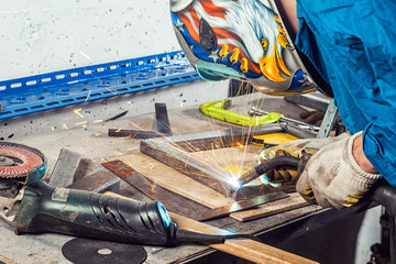 A man welder in a blue  overall, wearing a welding mask with an eagle and construction gloves, weld a metal welding machine in a workshop, sparks fly, instruments and an angle grinder  on the table