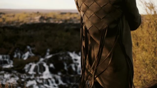 Close-up back view of young woman standing in front of Barnafoss waterfall in Iceland. Fringe from jacket waves on wind.
